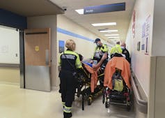 Paramedics wheeling a gurney past other paramedics and other gurneys in a hospital corridor