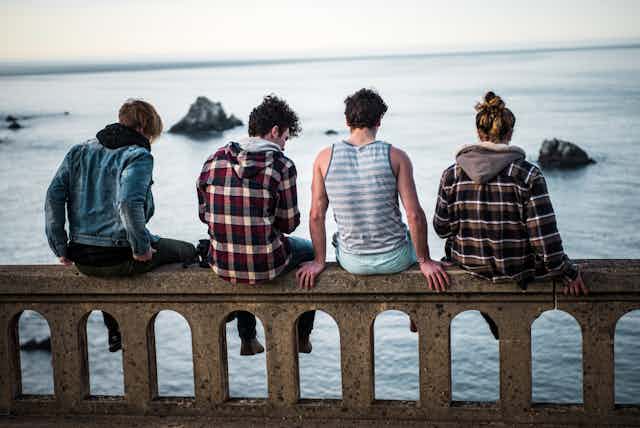 Un groupe de quatre jeunes, assis sur un muret et vus de dos, regarde l'océan.