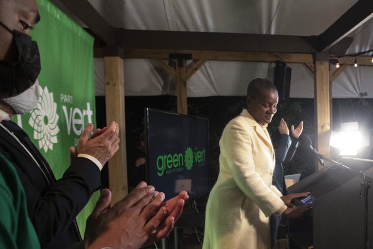 Annamie Paul in a cream suit speaks into a microphone at a podium as people behind her applaud.