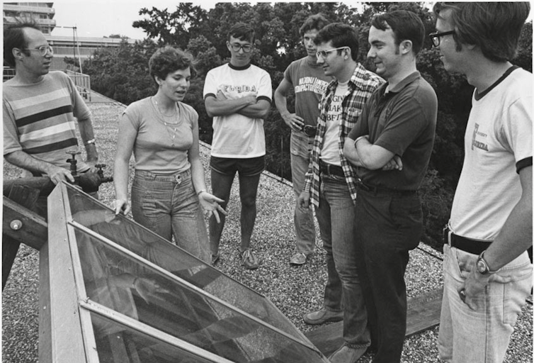 Black and white photo of a woman speaking to six men gathered around a solar panel.