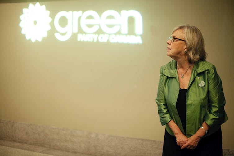May stands in front of a Green Party sign, wearing a green jacket.