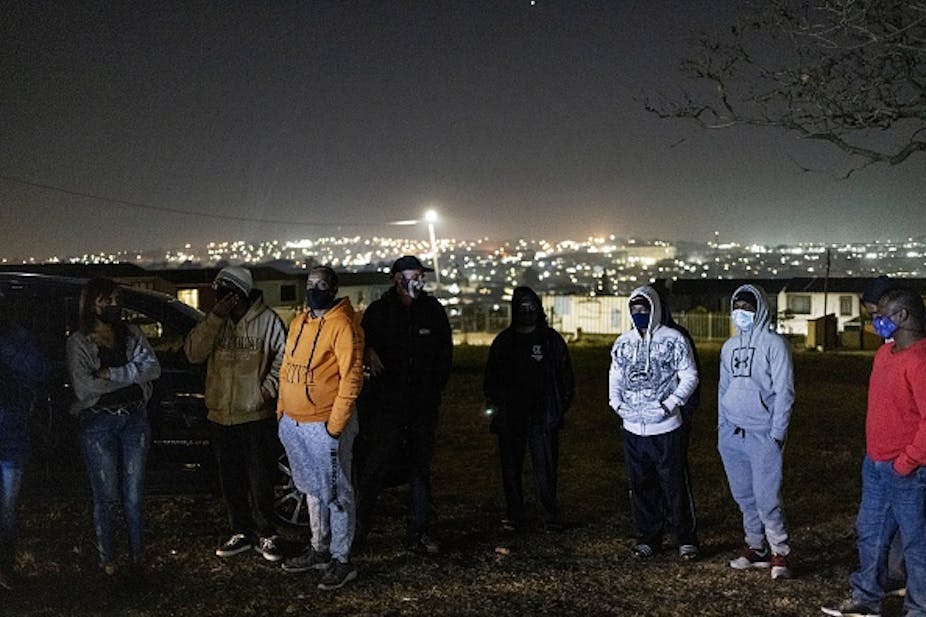 People wearing masks and warm clothing stand outdoors at night, with city lights in the background