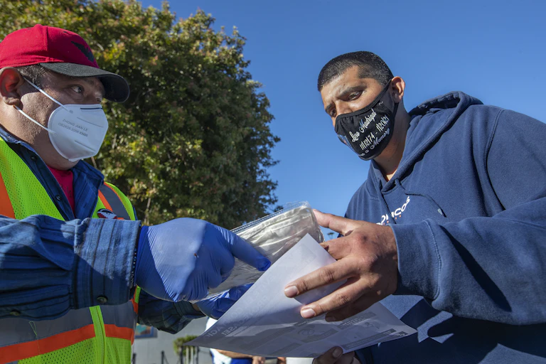 Man in red cap wearing gloves gives another man a packet of face masks