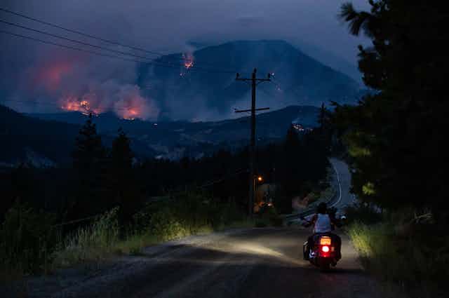 A man sits on his motorcycle on the side of the road, watching a wildfire burn
