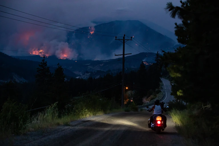 A man sits on his motorcycle on the side of the road, watching a wildfire burn