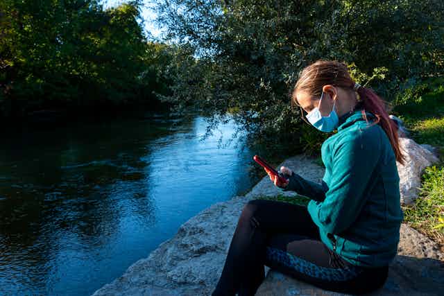 Adolescent girl with ponytail and wearing a mask sits beside a river, looking at her phone.