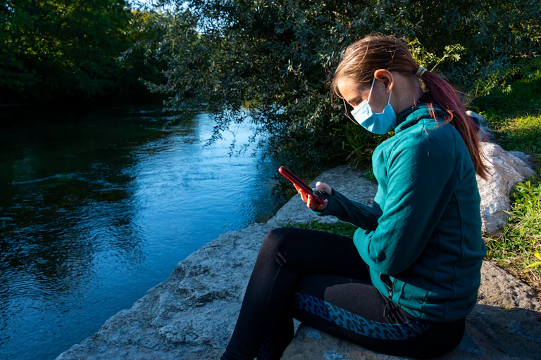 Adolescent girl with ponytail and wearing a mask sits beside a river, looking at her phone.