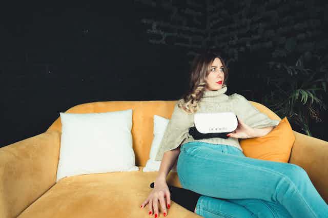 A woman with red lips and nails sits on a mustard couch and holds a VR headset