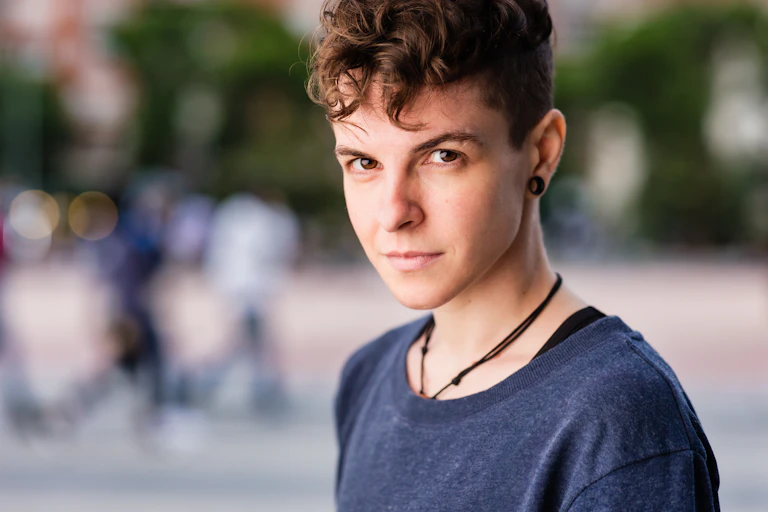 Young person with short brown hair, wearing a blue shirt stares at the camera, a small smile on their face.