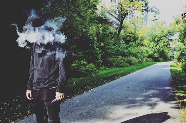 A man with his face obscured in a cloud of vaping smoke walks down a sidewalk.