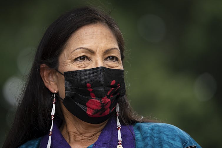 Secretary of the Interior Deb Haaland wears a black face mask with red handprint on it