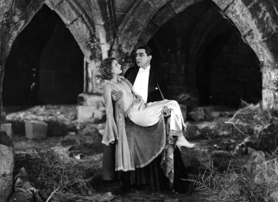 A vampire wearing white and black carries a woman in a gown amid castle ruins.