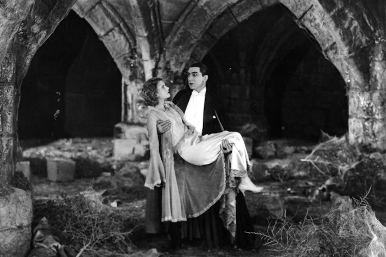 A vampire wearing white and black carries a woman in a gown amid castle ruins.