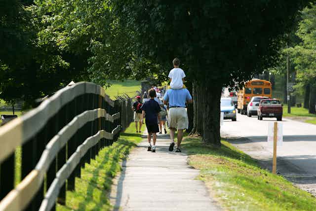 A man carries a child on his shoulders while walking along a scenic sidewalk with open space on one side and a school bus on the other.