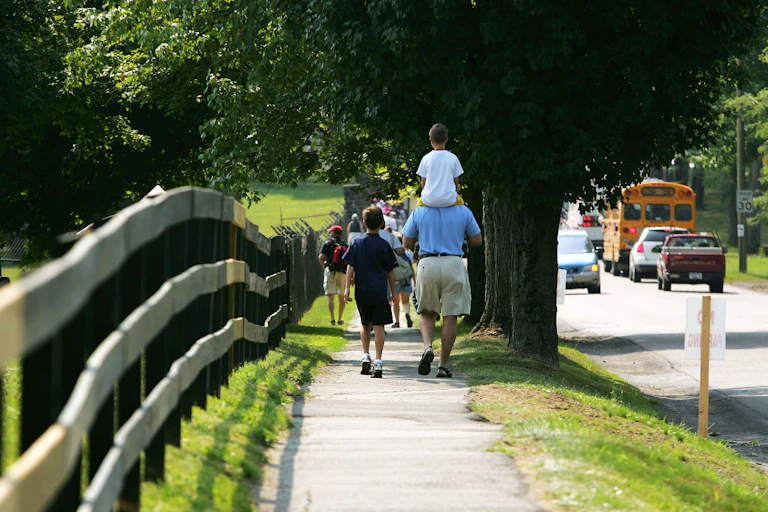 A man carries a child on his shoulders while walking along a scenic sidewalk with open space on one side and a school bus on the other.