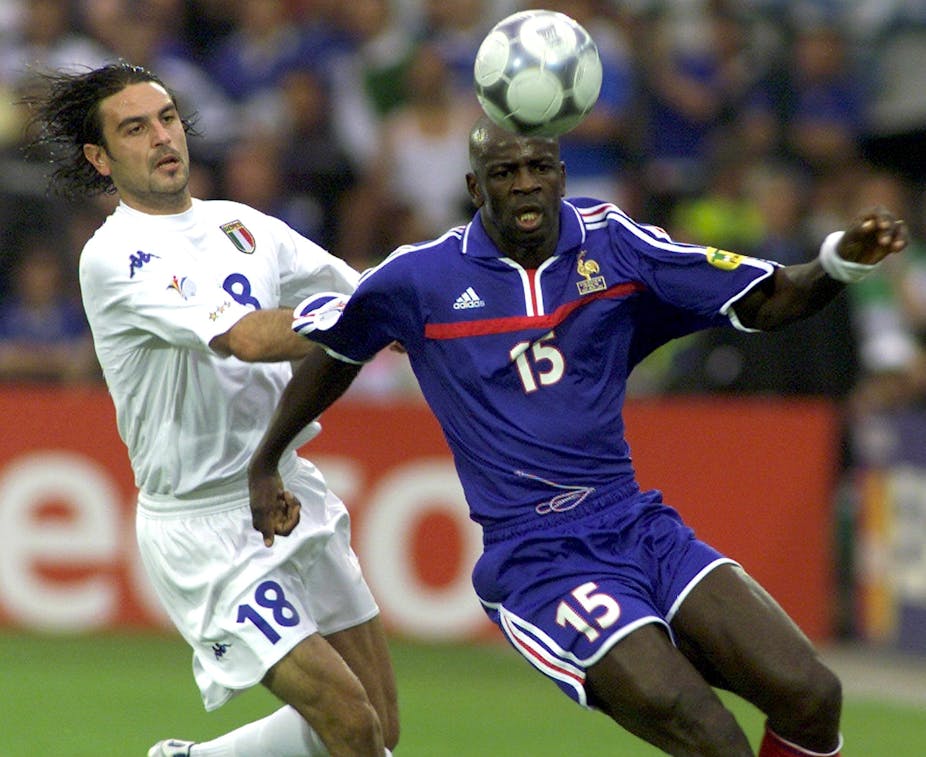 Two men, one dressed in white shorts and shirt and the other in blue, move towards a soccer ball