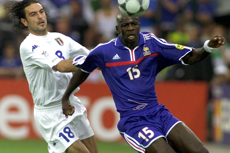 Two men, one dressed in white shorts and shirt and the other in blue, move towards a soccer ball