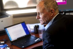 A man in a blue jacket sits at his desk with a laptop in front of him.