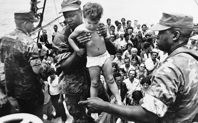 A U.S. Marine in uniform helps a Cuban toddler dressed only in a diaper off a boat.