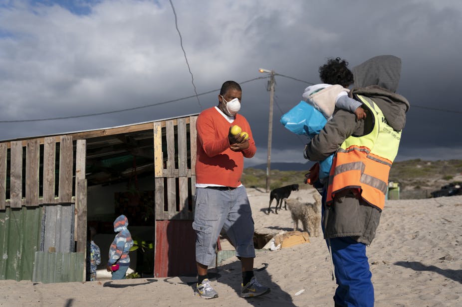 A man wearing a COVID-19 mask stands outside a shack, holding some apples. He is facing another man who is holding a child.
