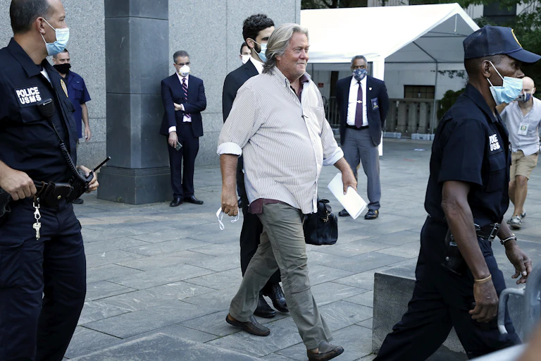 Steve Bannon in an untucked shirt and green slacks walsk from a courthouses surrounded by police officers.