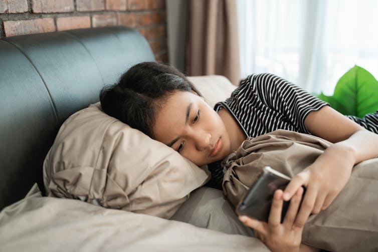 Young girl lays in bed, sad on her phone