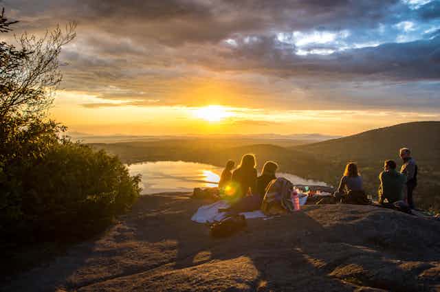 A group of people sit on a rocky outcropping on the top of a mountain overlooking a lake with the sun setting in the background