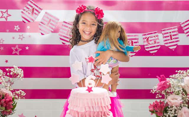 Young girls holds doll, with a pink cake in front and striped pink background behind her.