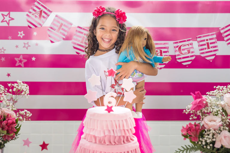Young girls holds doll, with a pink cake in front and striped pink background behind her.
