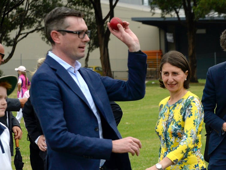 Man in suit jacket prepares to bowl a cricket ball watched by a woman