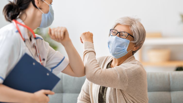 A nurse and an older woman, both wearing face masks, touching elbows in greeting