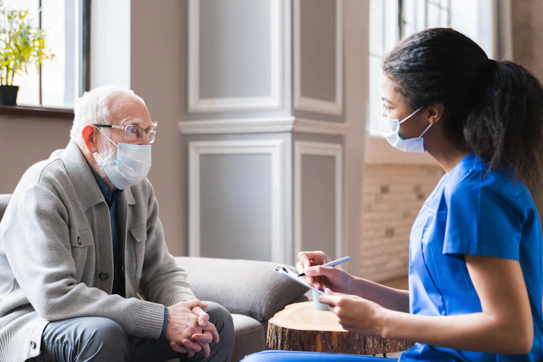 A care worker writes on a clipboard while speaking to an elderly man. Both are wearing face masks.