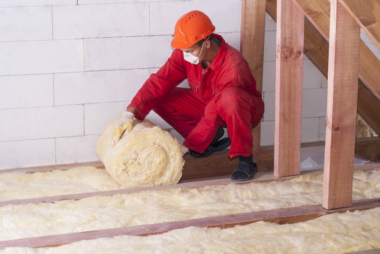 A worker in red overalls unrolls thermal insulation in a loft.