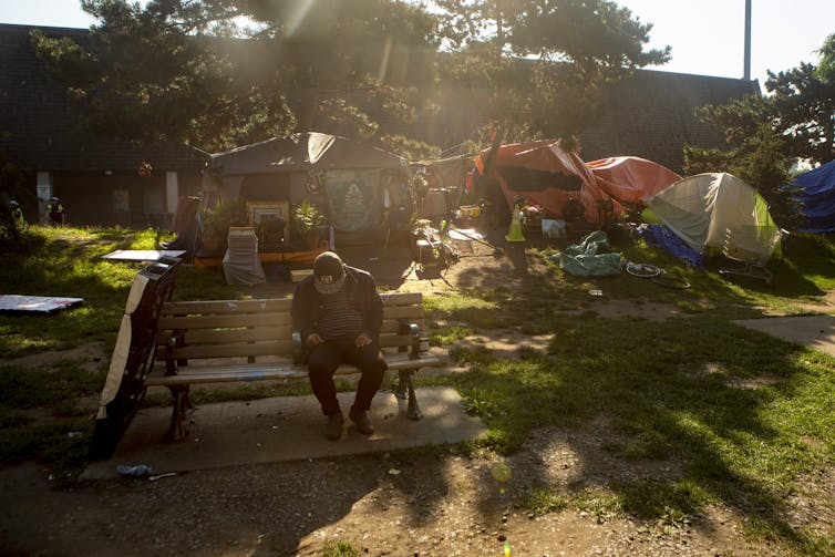A man sits on a bench in front of a homeless tent camp