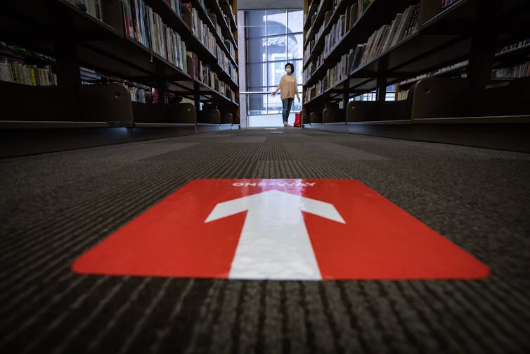 A big red arrow is shown in the foreground. A woman wears a face mask and plastic gloves while browsing books at the Vancouver Public Library's central branch.