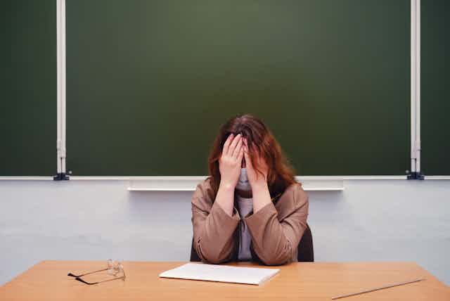 A while female teacher sits at her desk while she covers her head with her hands.