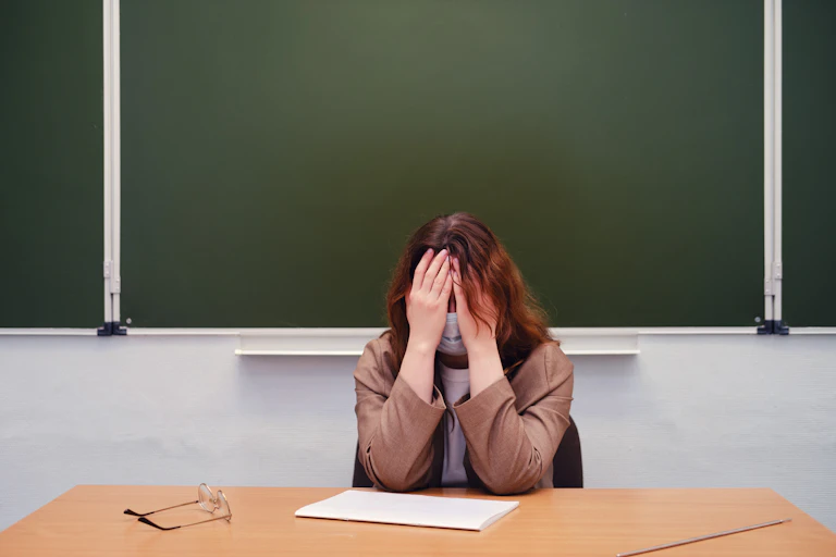 A while female teacher sits at her desk while she covers her head with her hands.
