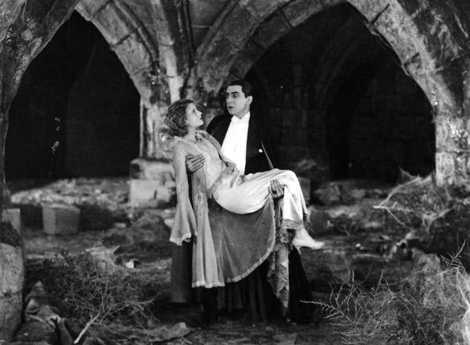 A vampire wearing white and black carries a woman in a gown amid castle ruins.
