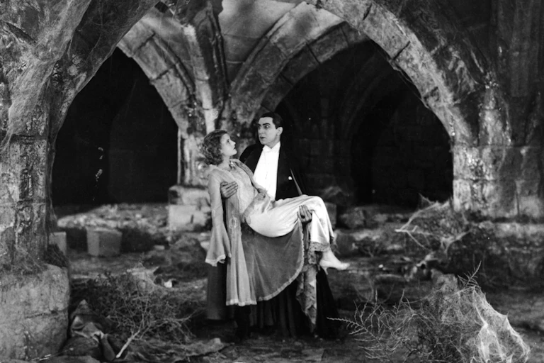 A vampire wearing white and black carries a woman in a gown amid castle ruins.