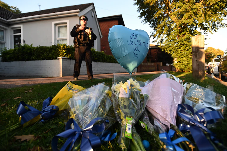 Flowers laid in Essex in memory of David Amess. A balloon reads 'RIP, we will miss you' and a police officer stands in the background.