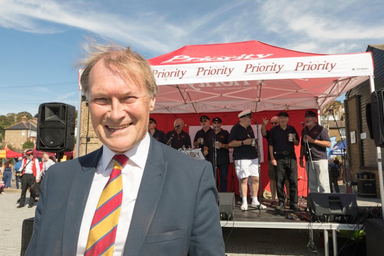 David Amess na frente de um estande de banda em seu distrito eleitoral em um dia de verão.