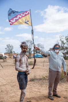 An Aboriginal person painted up, stands under a flag.