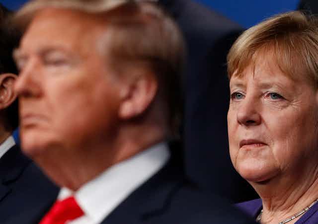 Former German Chancellor Angela Merkel looks at Donald Trump, seated in front of her. Trump is out of focus, while Merkel's face is in focus.