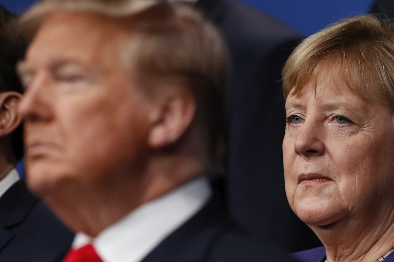 Former German Chancellor Angela Merkel looks at Donald Trump, seated in front of her. Trump is out of focus, while Merkel's face is in focus.
