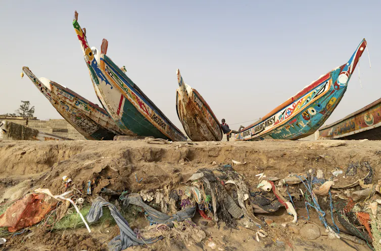 Plastic waste eroding out of beach, above stands a boy between painted boats