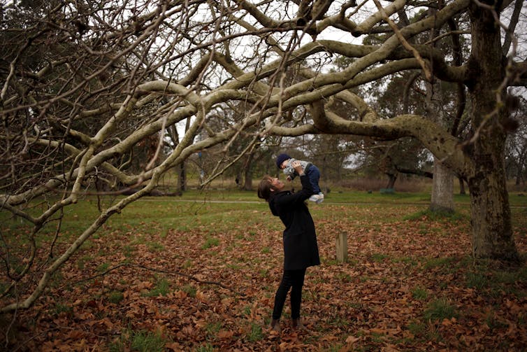 A mother in a park lifts her baby over her head.