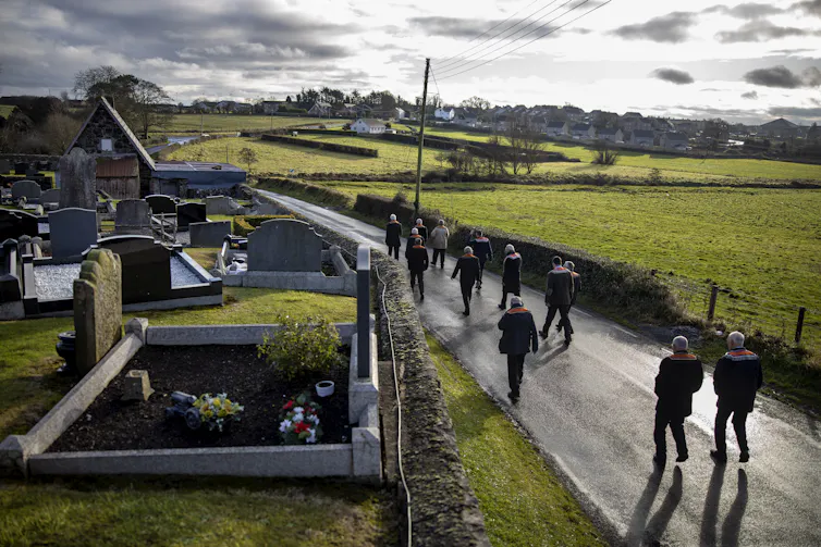 A handful of men with their backs to the camera walk down a country road, with a cemetery to their left and fields to their right.