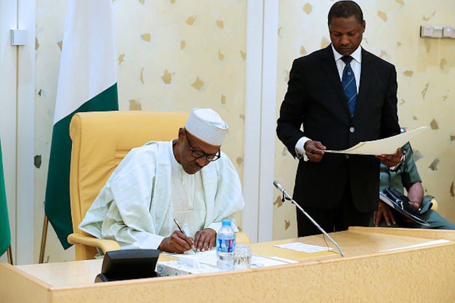 A man wearing a cap writing on a papers and another man standing beside him.