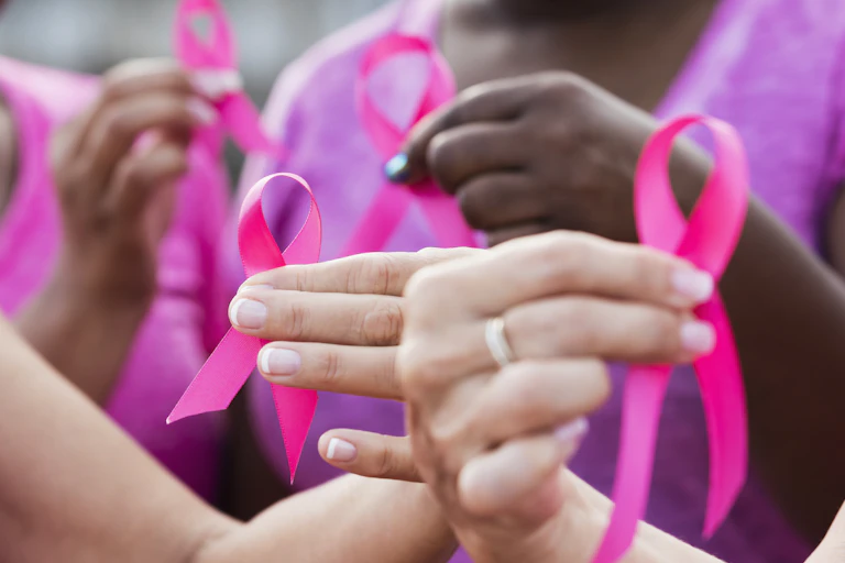 four multi-ethnic women wearing pink shirts, holding breast cancer awareness ribbons. The focus is on one of the hands.