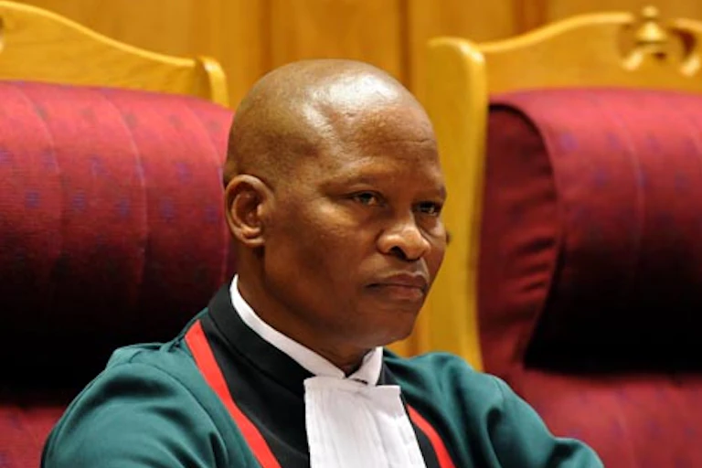 A bald-headed, clean-shaven man dressed in legal garb sits on a chair while preciding over court proceedings
